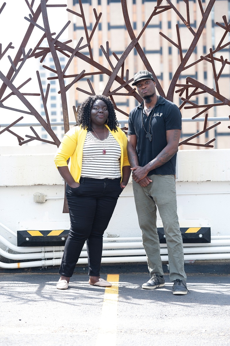 A Black woman wearing a yellow cardigan, striped t-shirt and black jeans and a beared Black Man wearing a black monogrammed shirt and khaki pants.