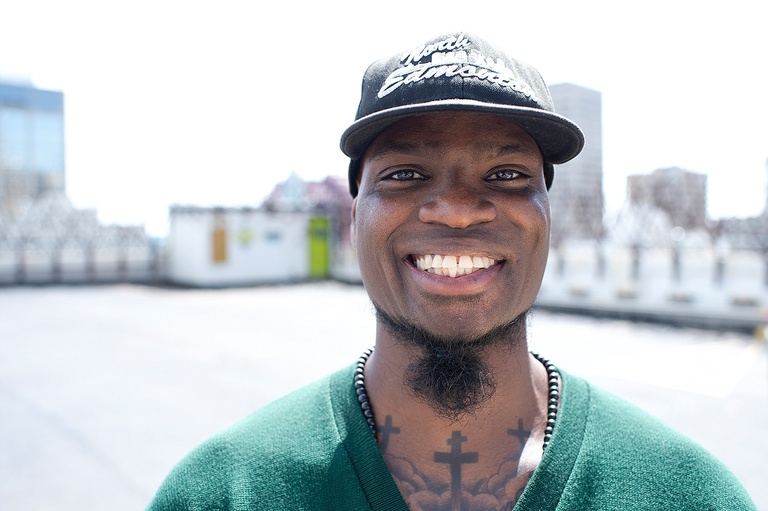 Bearded Black man with a smile. His chest tattoos are visible as he is wearing a v-neck green sweater.